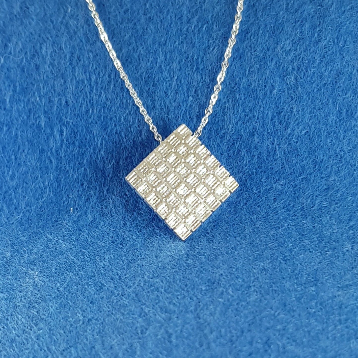 Square pendant which hangs from the corner with cross stitch aida fabric texture and dainty 18 inch chain in sterling silver.